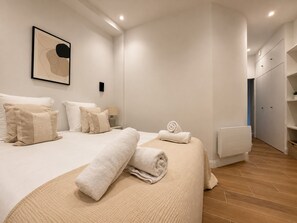 A modern bedroom with a bed, pillows, a rolled towel, a bedside table with a lamp, and a wall-mounted artwork.