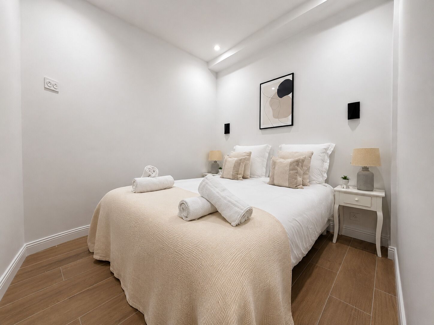 A bedroom with a large bed, two bedside tables, a wall-mounted lamp, and a framed artwork.