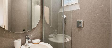 A modern bathroom with a glass-enclosed shower, a round mirror, and a white sink with a cup and soap dish.