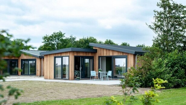 Een modern huis met twee verdiepingen, grote glazen deuren en ramen, een overdekt terras met tuinmeubilair en een verzorgd gazon.