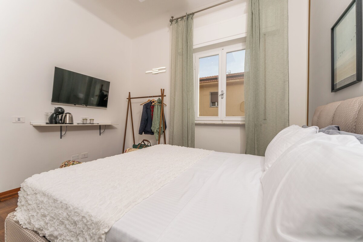 Quarto com cama, televisão na parede, janela com cortinas e prateleira com objetos.