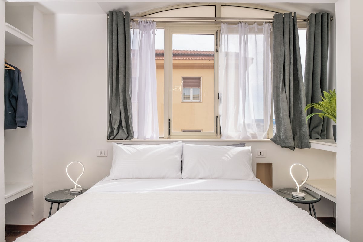 Quarto com cama, duas mesas de cabeceira, janela com cortinas e vista exterior.