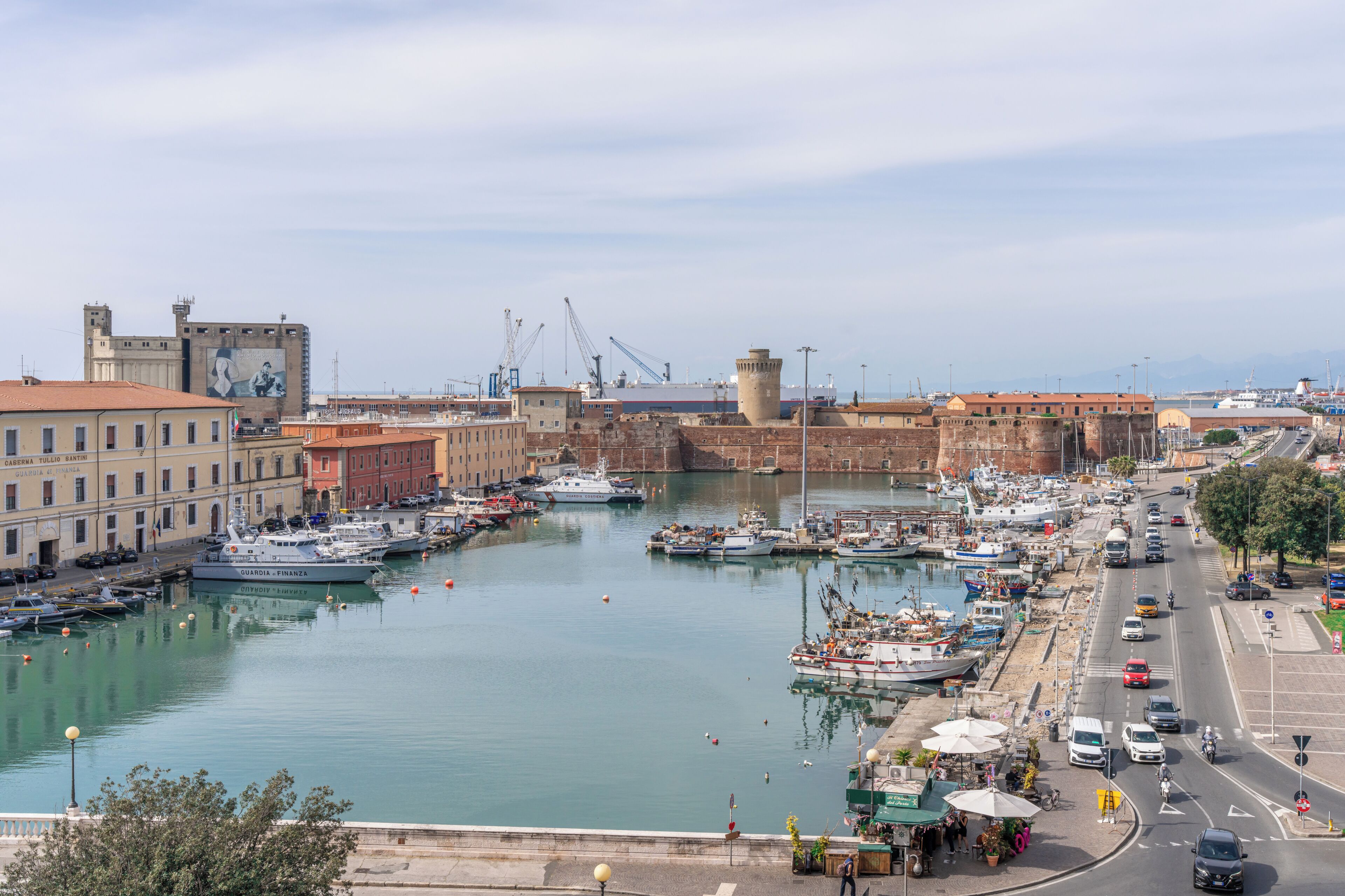 A marina with boats docked, a historic fort, and a busy street with cars and a café.