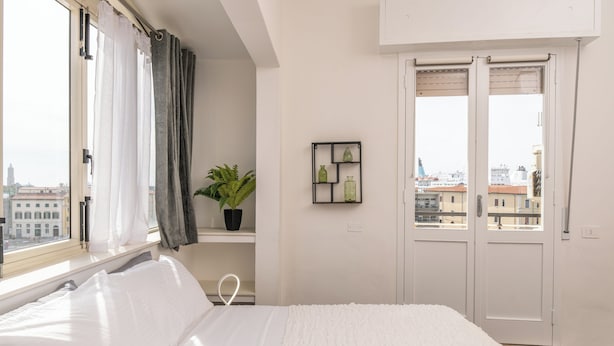 A bedroom with a bed, a window with curtains, a potted plant, and a door with a view of buildings outside. - Porto Mediceo (Livorno)
