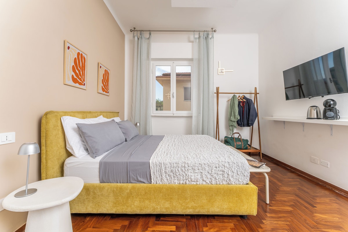 Quarto moderno com cama, mesa de cabeceira, luminária e televisão na parede.