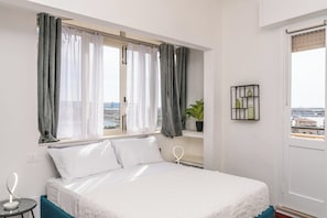 Ein Schlafzimmer mit Bett, Nachttisch mit Lampe, einem Fenster mit Vorhängen und Blick auf die Stadt.