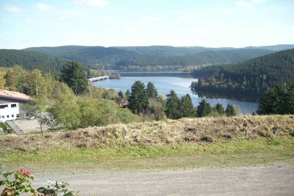 Ein See, der von bewaldeten Hügeln umgeben ist, mit einer Brücke und einem Gebäude mit rotem Dach.