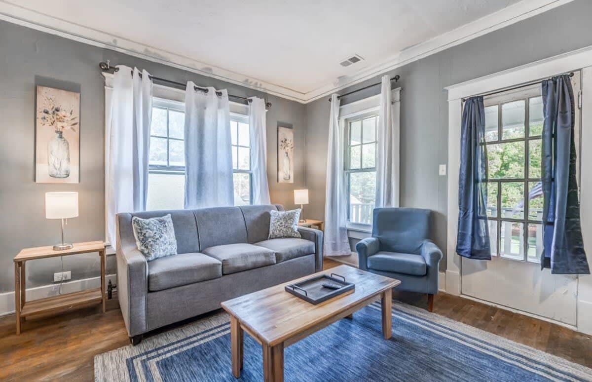 A living room with a grey sofa, a blue armchair, a wooden coffee table, and a blue rug. There are two windows with curtains, a lamp on a side table, and artwork on the walls.