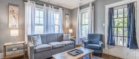 A living room with a grey sofa, a blue armchair, a wooden coffee table, and a blue rug. There are two windows with curtains, a lamp on a side table, and artwork on the walls.