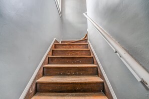 Eine schmale Treppe mit Holzstufen und weißen Geländern, die in einem Korridor mit grauen Wänden nach oben führt.