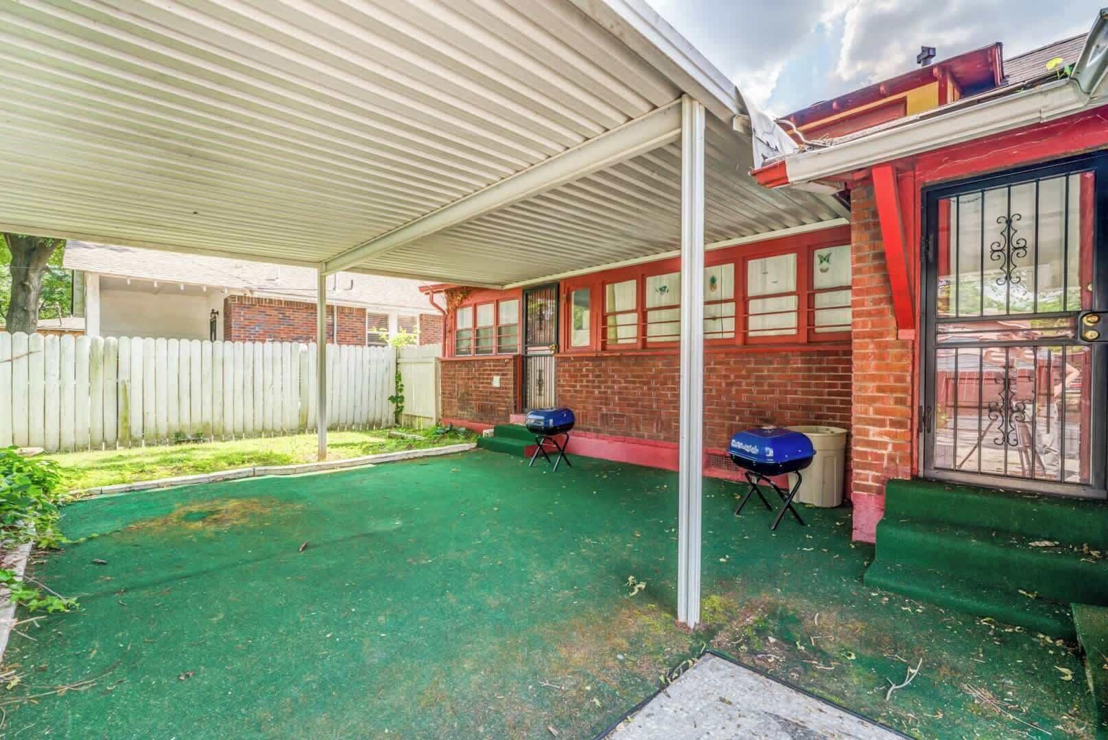 A covered outdoor area with a green artificial turf, two barbecue grills, a white fence, and a brick building with windows.