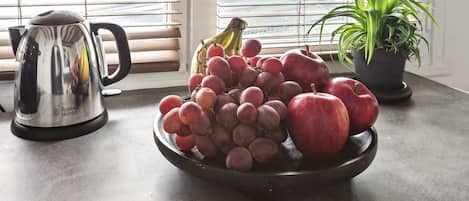 Una encimera de cocina con un frutero que contiene uvas, manzanas y un plátano, una tetera y una planta en maceta.