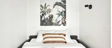 Un lit bien fait, avec du linge de lit blanc et des coussins à motifs, un petit tapis et une pile de serviettes pliées posées sur le sol.