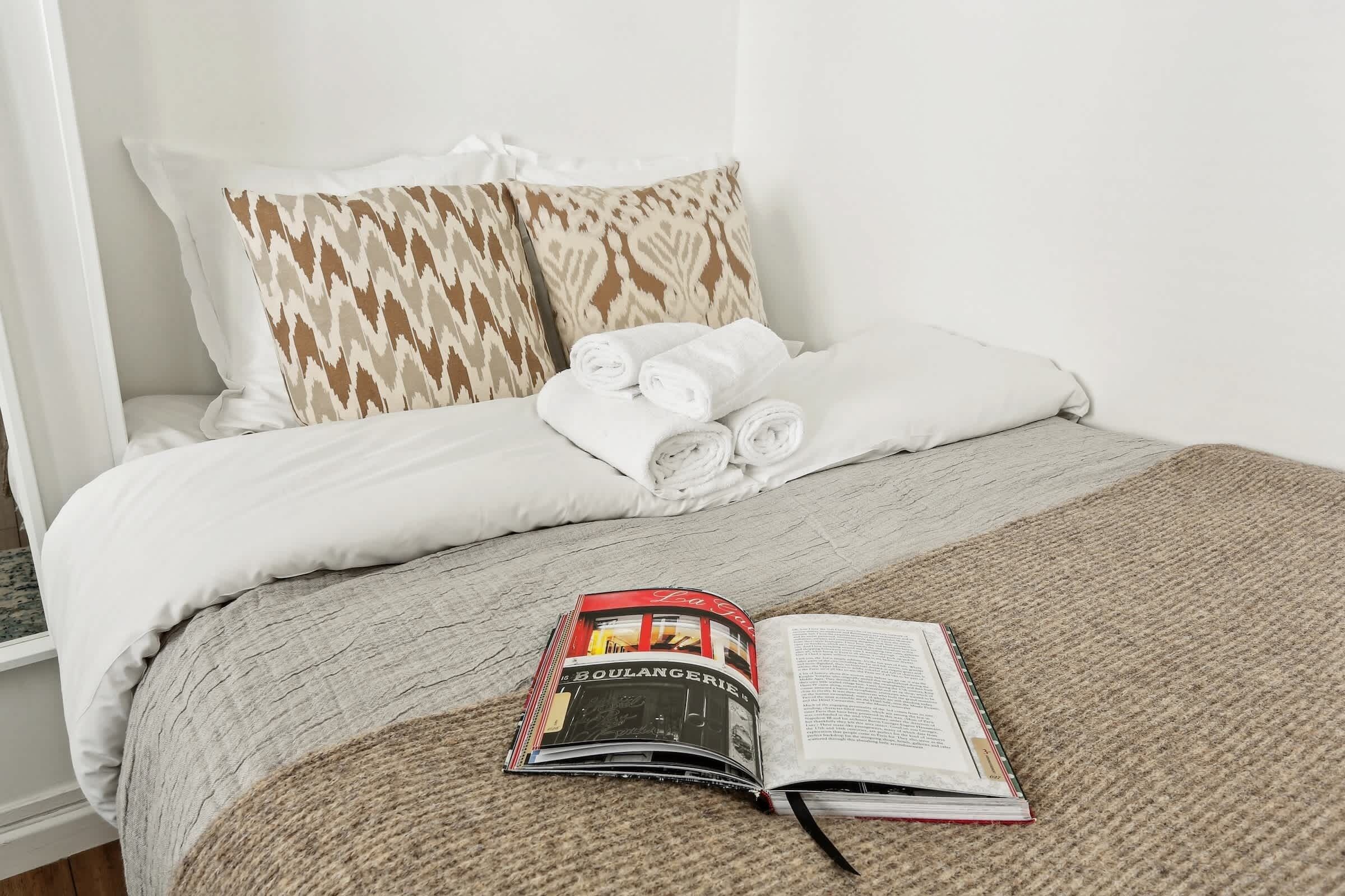 A neatly made bed with white and patterned pillows, a rolled towel, and a book on a beige blanket.