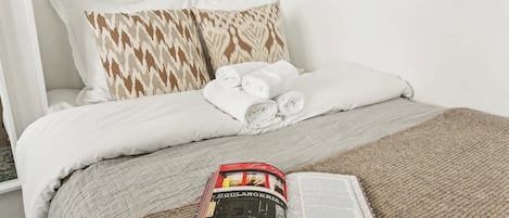 A neatly made bed with white and patterned pillows, a rolled towel, and a book on a beige blanket.