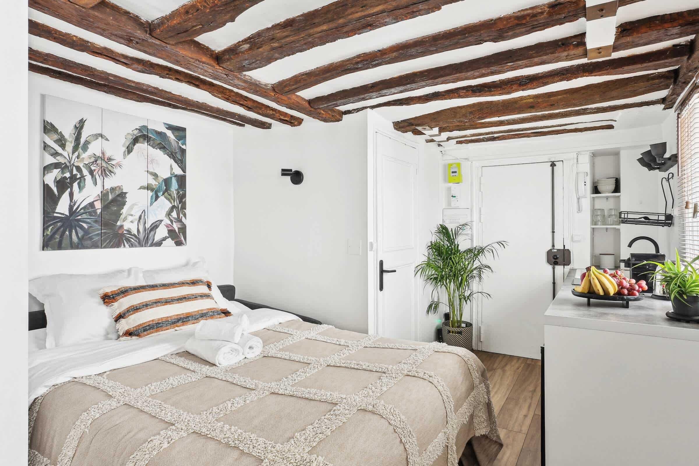 A bedroom with a bed, a nightstand, a potted plant, and a view into the kitchen.