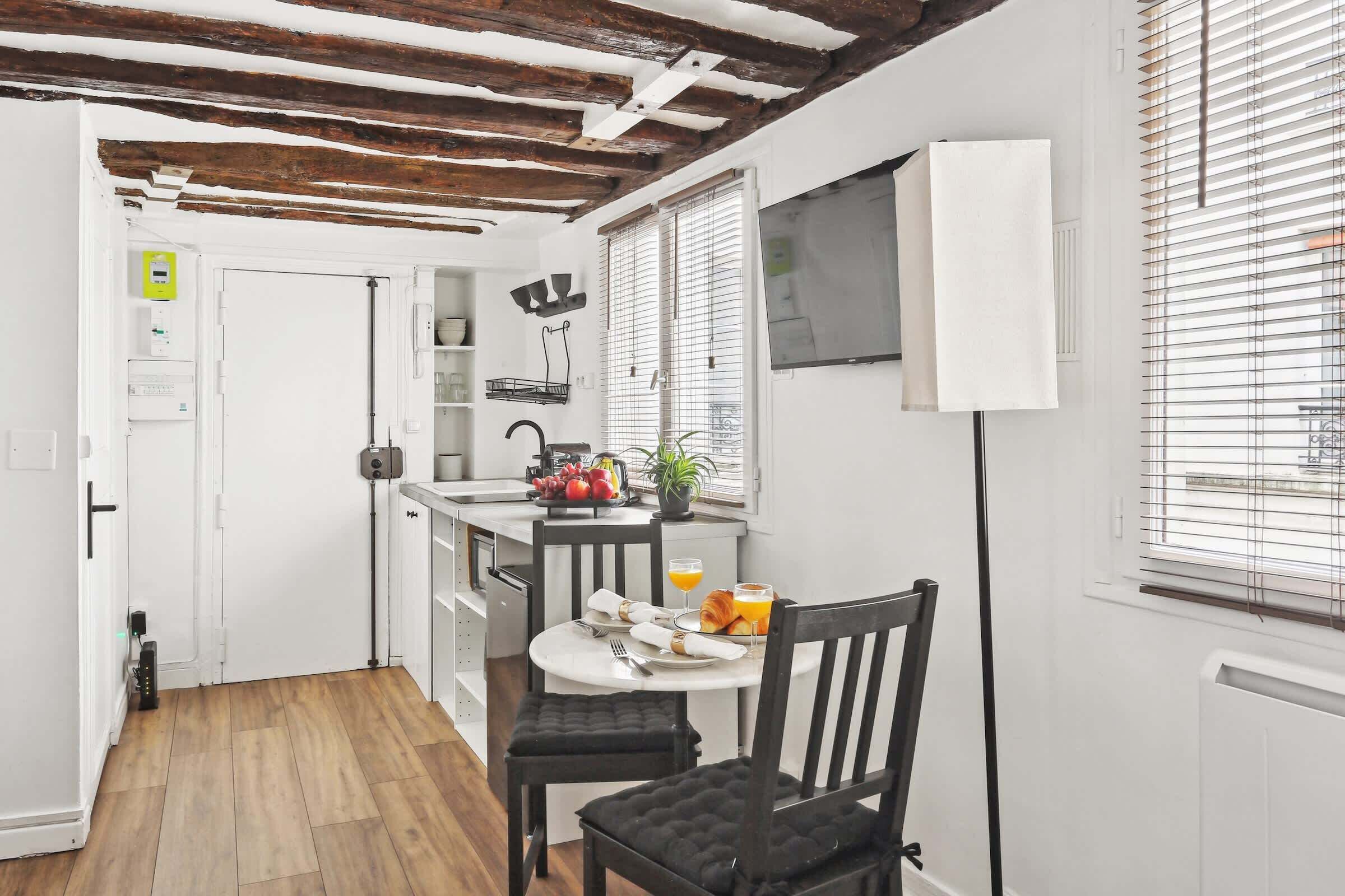 A compact kitchen with a dining area, wooden ceiling beams, and a view of the street through the window.