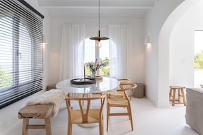 Een eetgedeelte met een ronde tafel, houten stoelen, een bank en uitzicht op groen door grote ramen.