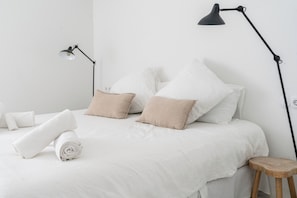 Een net opgemaakt bed met witte beddengoed en twee beige kussens, een zwarte vloerlamp en een houten kruk.