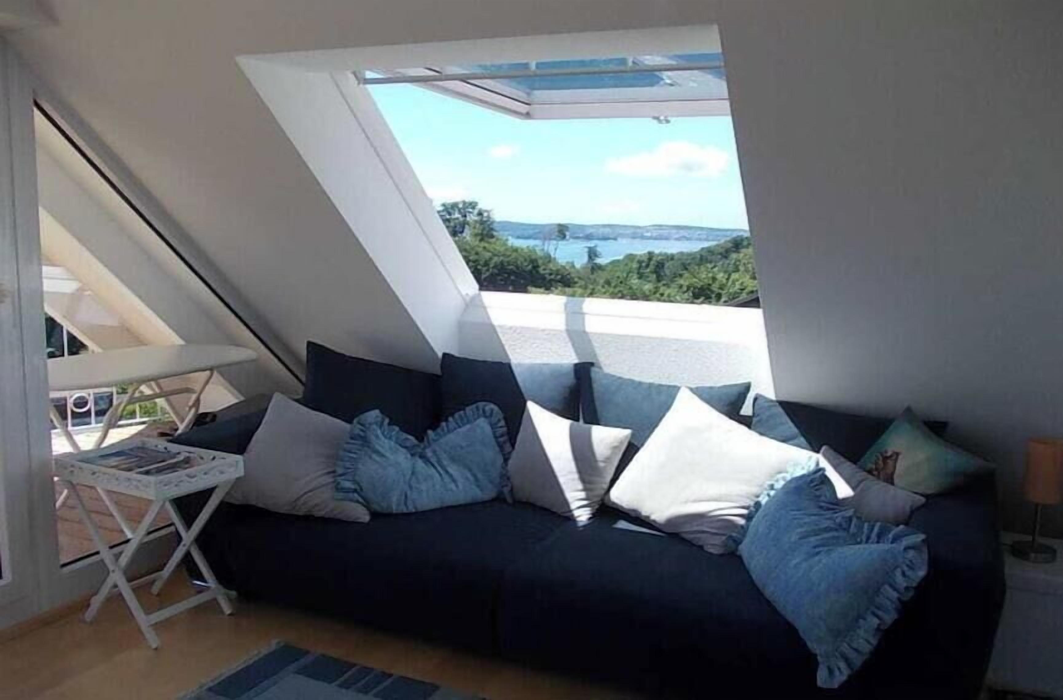 A room with a dark-colored sofa, a small table with a magazine, and a view of a body of water through a skylight.