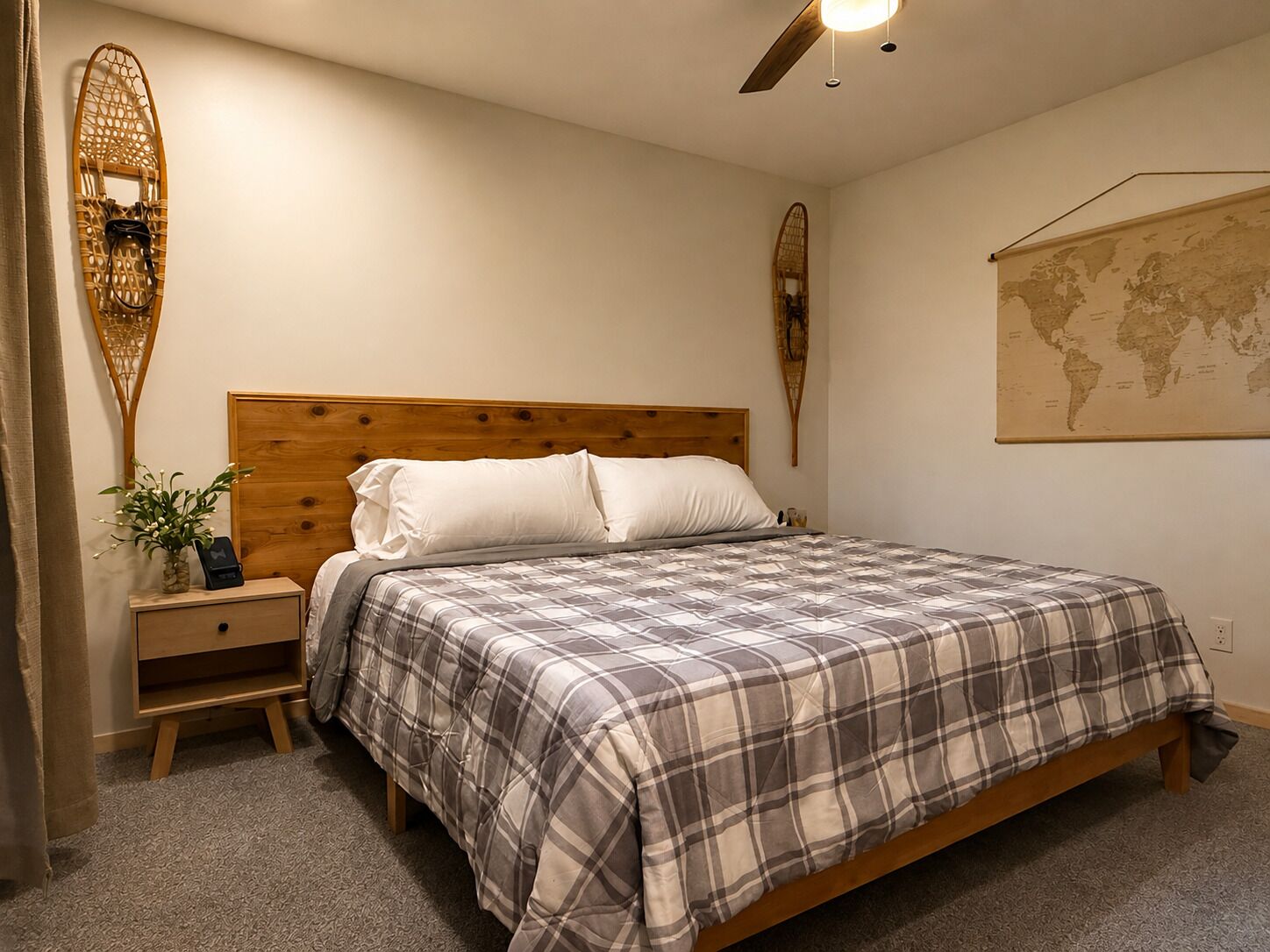 A bedroom with a wooden headboard, a bed with a plaid bedspread, a nightstand with a vase of flowers, and a world map on the wall.