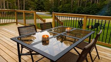 A glass-top table with chairs on a wooden deck, a candle on the table, and a view of a forested area.