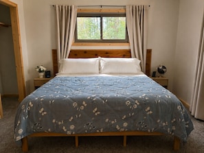 A bedroom with a wooden bed, a window with curtains, a bedside table with a globe and a plant, and a nightstand with a clock. - Wooded Retreat Near Wisconsin Dells + EV Charging - Short Walk to Winery (Wisconsin Dells)