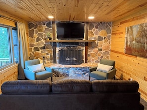Una sala de estar acogedora con chimenea de piedra, dos sillones y un televisor grande montado sobre la chimenea.