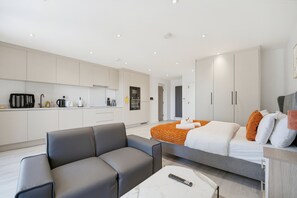 Ein modernes Hotelzimmer mit einem grauen Sofa, einem Bett mit orangefarbener Decke, einem weißen Couchtisch und einer Küchenzeile im Hintergrund.