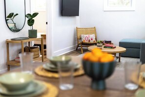 A modern living room with a dining table set, a television mounted on the wall, and a comfortable seating area. - Quiet Artful Cottage · Steps to Ocean View Beach (Norfolk)