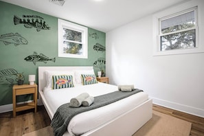 A bedroom with a bed, pillows, a nightstand, a window, and fish-themed wall art. - Quiet Artful Cottage · Steps to Ocean View Beach (Norfolk)