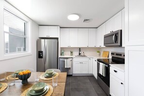 A modern kitchen with white cabinetry, stainless steel appliances, and a dining area with a wooden table and placemats. - Quiet Artful Cottage · Steps to Ocean View Beach (Norfolk)