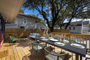 An outdoor deck with a dining table set for six, surrounded by wicker chairs and a wooden railing. - Quiet Artful Cottage · Steps to Ocean View Beach (Norfolk)