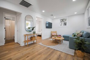 A living room with a sofa, a coffee table, a chair, a TV, and a mirror. - Quiet Artful Cottage · Steps to Ocean View Beach (Norfolk)