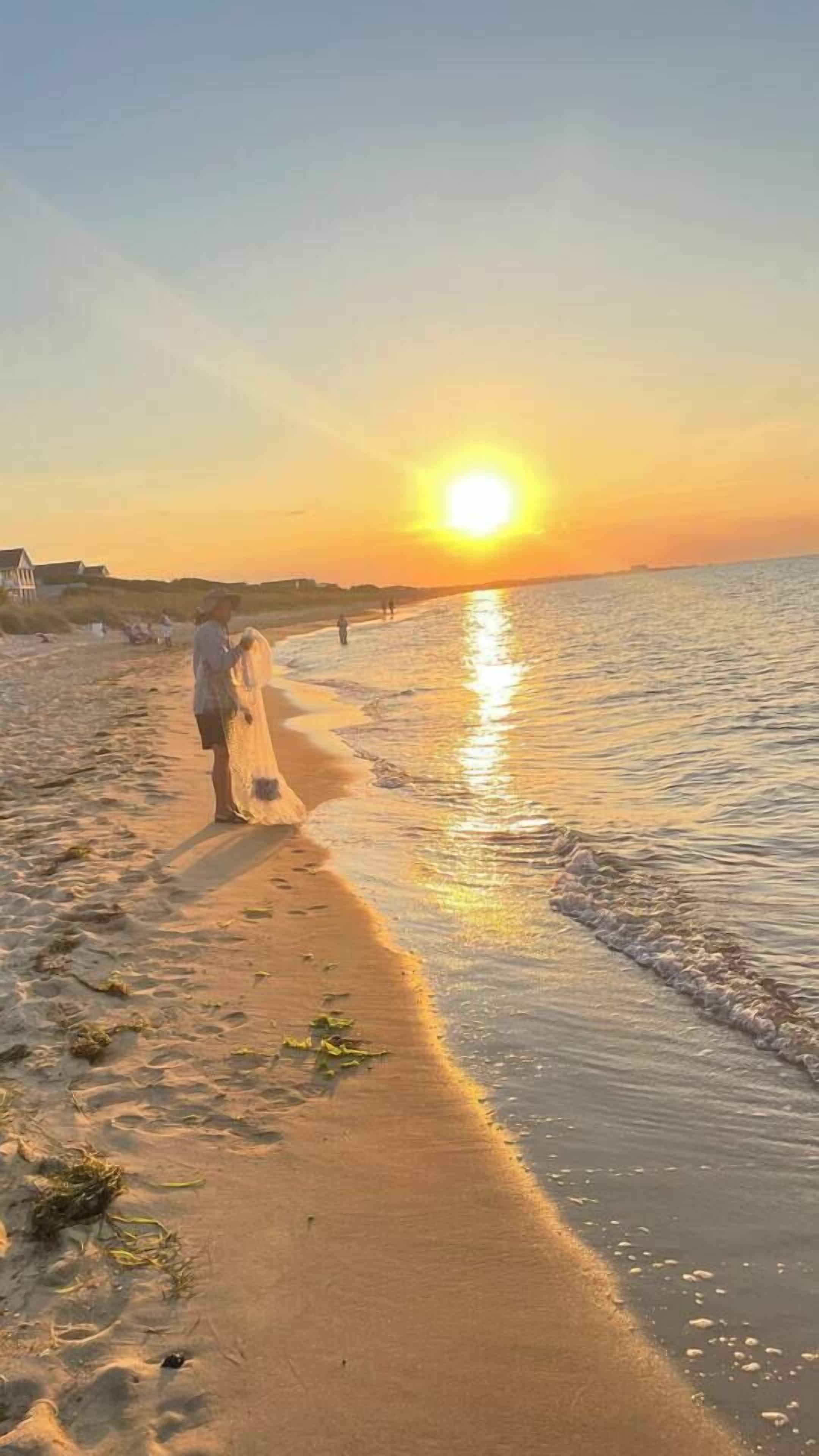 Eine Strandansicht mit Sonnenuntergang, ein Paar geht am Ufer entlang und ein Gebäude im Hintergrund.