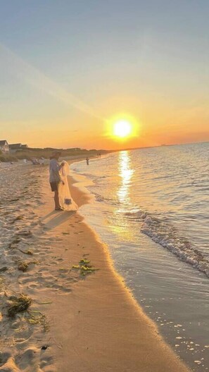 A beach scene with a sunset, a couple walking along the shore, and a building in the background. - Quiet Artful Cottage · Steps to Ocean View Beach (Norfolk)