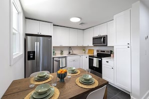 A modern kitchen with white cabinets, stainless steel appliances, and a wooden dining table set with plates and glasses. - Quiet Artful Cottage · Steps to Ocean View Beach (Norfolk)