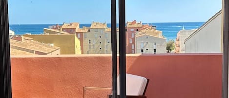 A balcony with a wooden bench and a chair, overlooking a coastal cityscape with buildings and the sea.