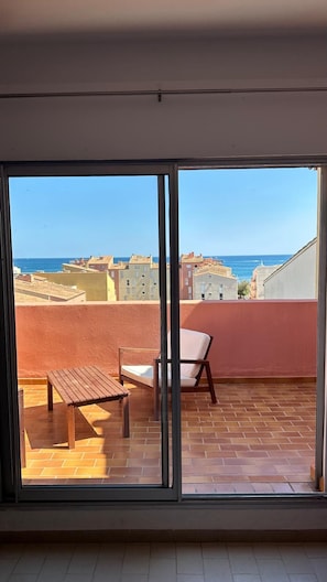 Un balcón con un banco de madera y una silla, con vistas a un paisaje urbano costero con edificios y el mar.