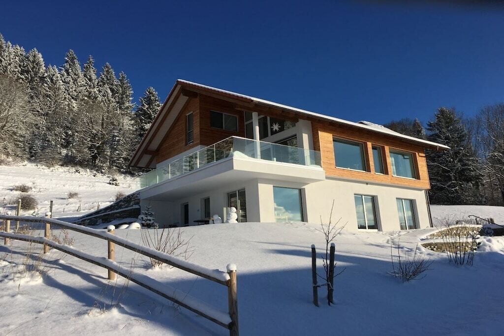 Una casa moderna de dos pisos con un balcón, rodeada de árboles cubiertos de nieve.