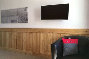 A room with a wooden panel wall, a black leather chair with a red and grey cushion, and a black flat-screen TV mounted on the wall. - Holiday apartment with 2 bedrooms (Füssen)