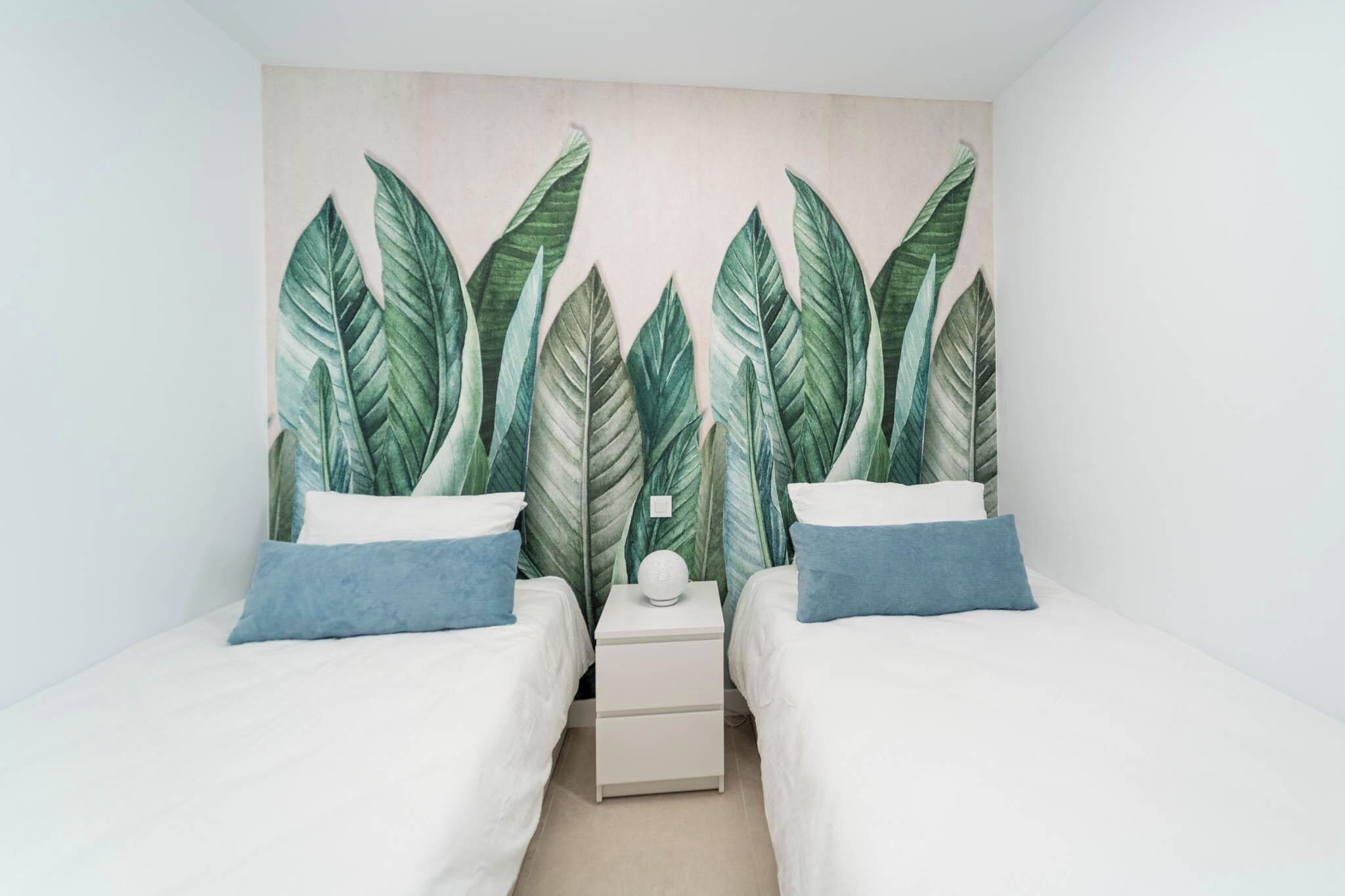 Une chambre avec deux lits, du linge de lit blanc et une fresque murale représentant des feuilles vertes.