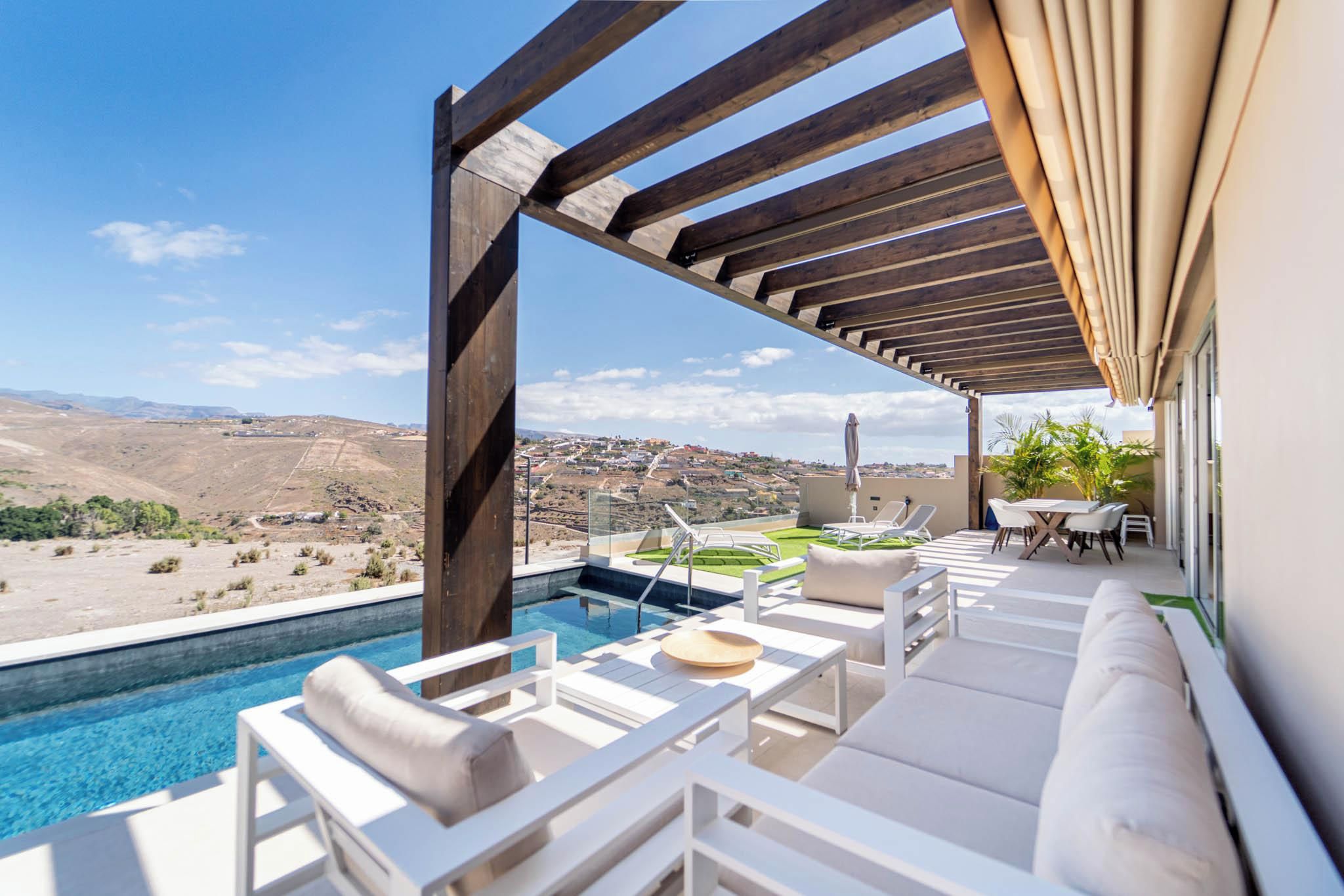 A poolside area with a view of a hilly landscape, outdoor seating, and a pergola structure.