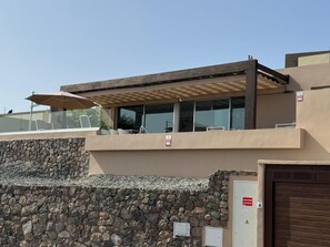 A modern building with a stone retaining wall, a covered outdoor seating area, and a gated entrance. - Kaiman 19 by VillaGranCanaria (San Bartolomé de Tirajana)