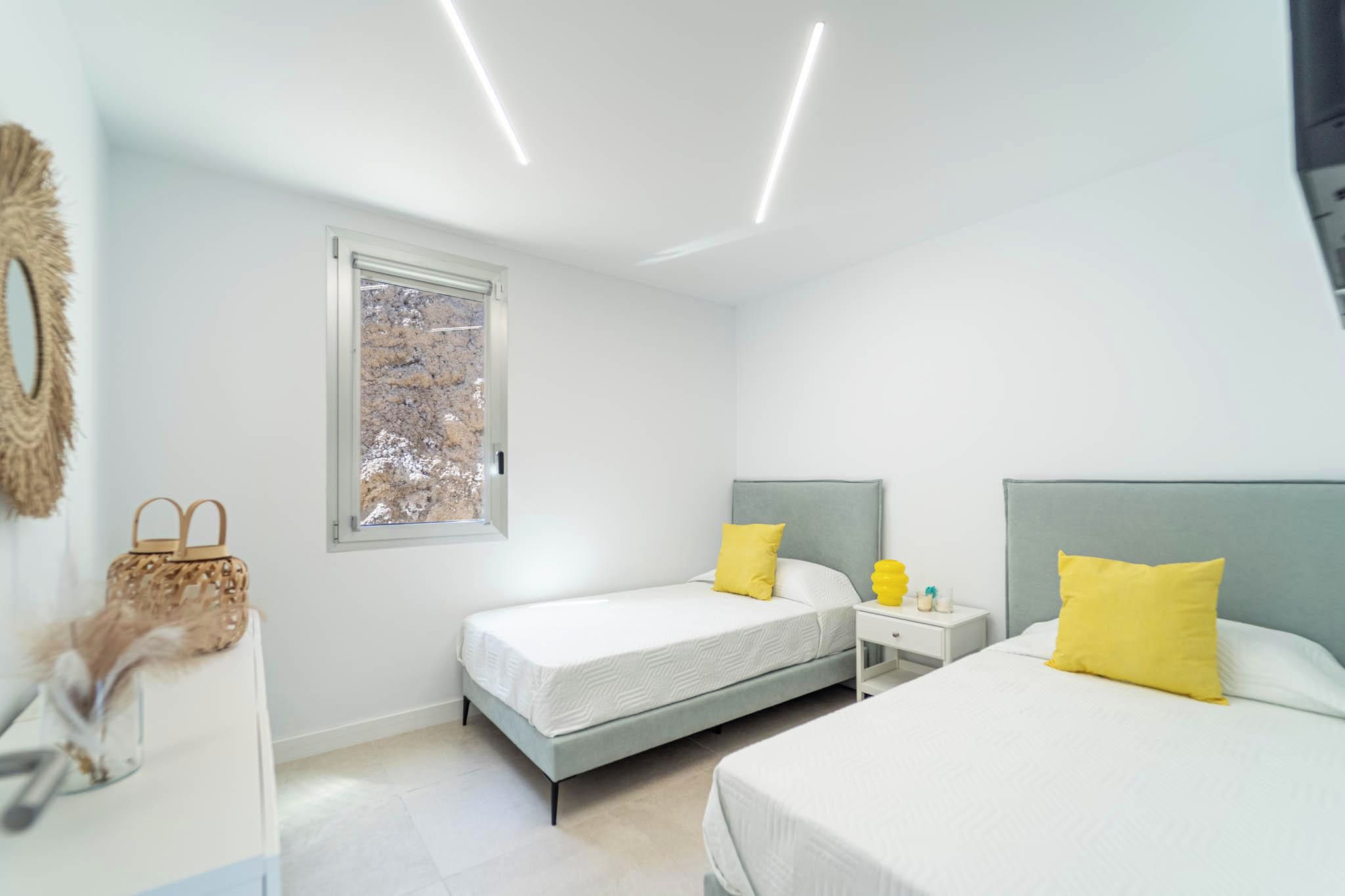 A modern bedroom with two beds, a window, and a bedside table with a lamp.