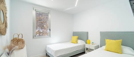 A modern bedroom with two beds, a window, and a bedside table with a lamp.