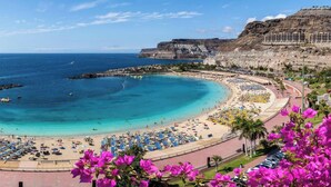 Una playa costera con una línea de costa curva, un paseo marítimo y un edificio al borde de un acantilado.