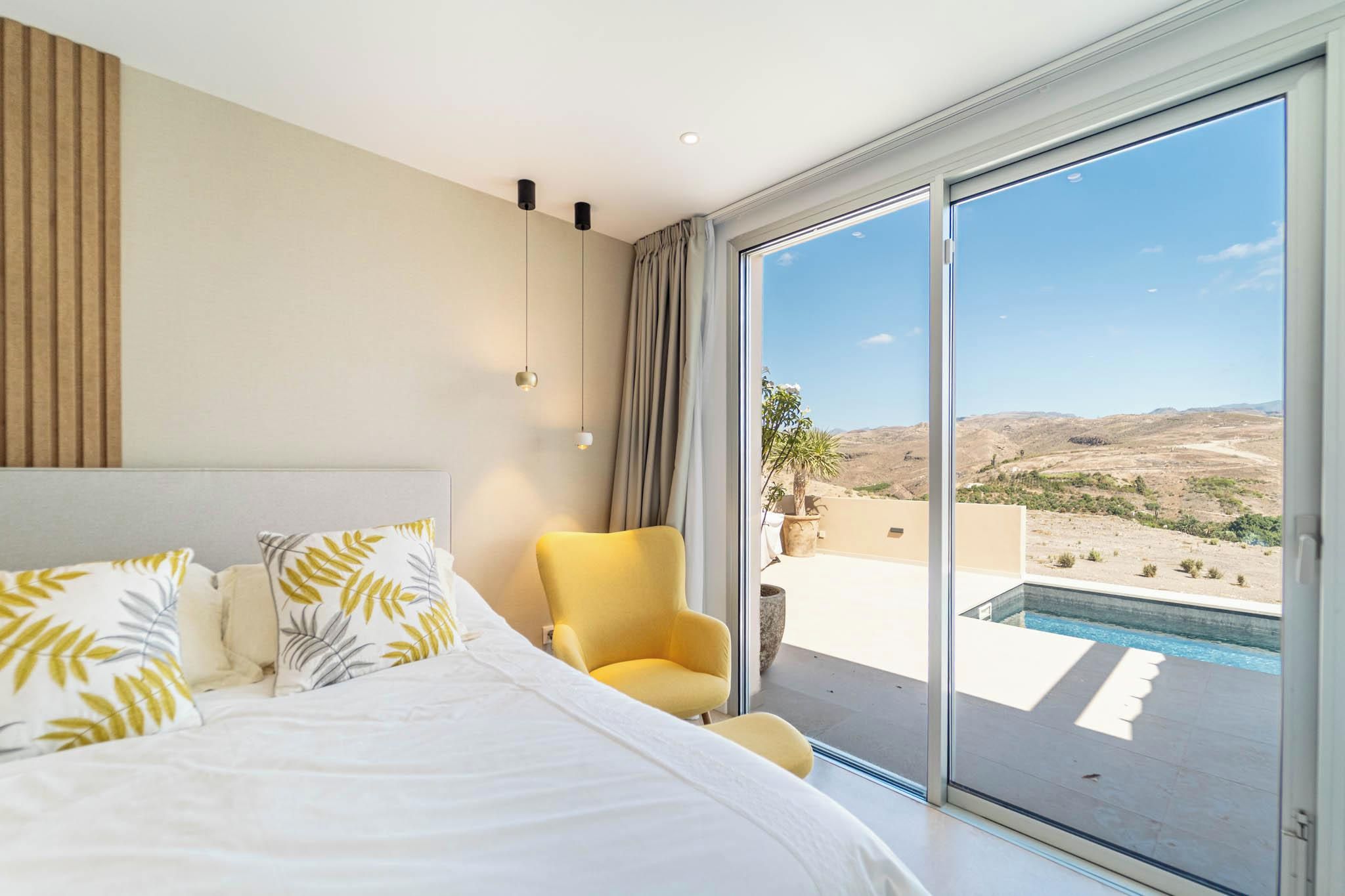 Un dormitorio con una cama grande, un sillón amarillo y vistas a una piscina y al paisaje.