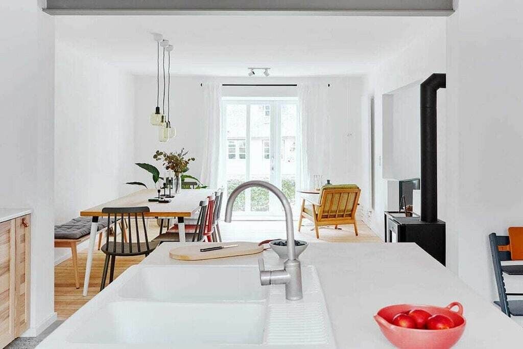 Et moderne køkken med en central ø, et spiseområde med et træbord og stole, og udsigt udad gennem et stort vindue.
