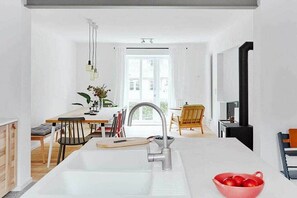 A modern kitchen with a central island, a dining area with a wooden table and chairs, and a view of the outside through a large window. - Holnis Customs House (Glücksburg)